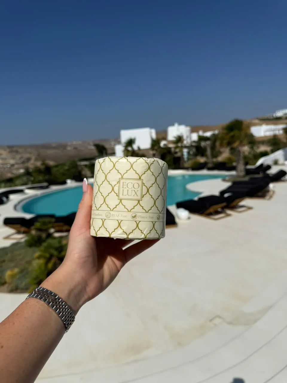 A hand holding a wrapped Eco Lux roll above a pool terrace with whitewashed houses and sea