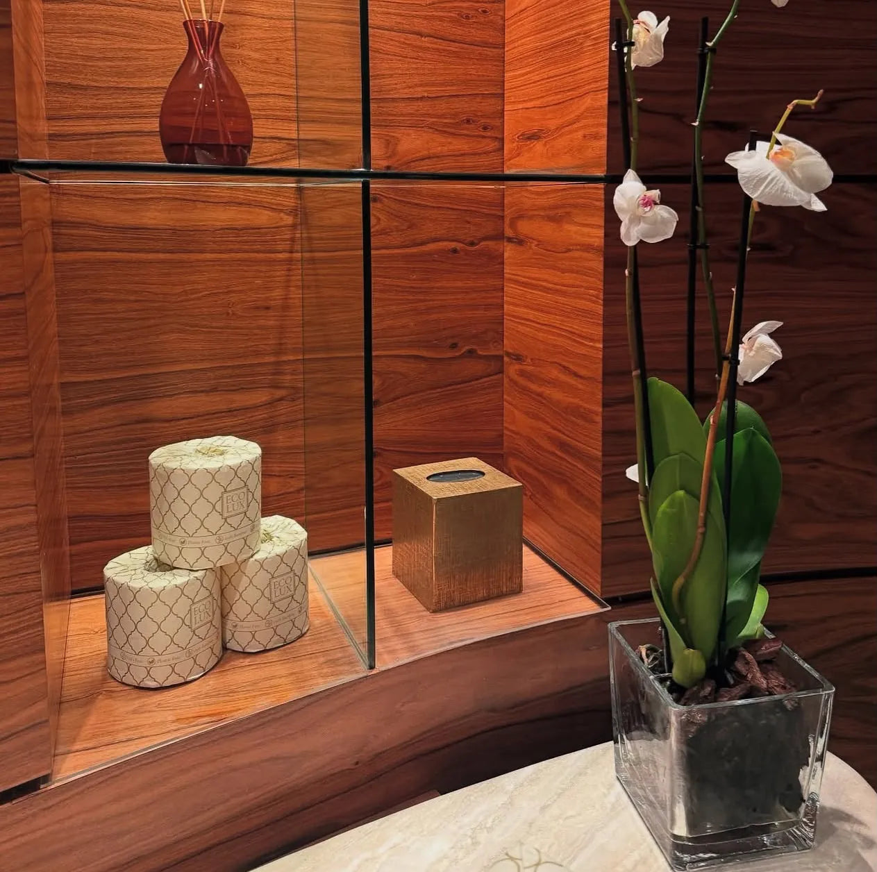 Three wrapped Eco Lux rolls in a lit walnut wall niche beside a white orchid